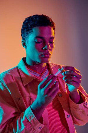 Afrikanisch-amerikanischer Kerl schaut auf Zigarettenrolle auf buntem Hintergrund mit blauer Neonbeleuchtung