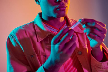 beschnitten afrikanisch-amerikanischer Kerl hält gedrehte Zigarette auf buntem Hintergrund mit blauer Neonbeleuchtung