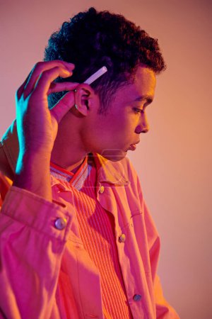 Téléchargez les photos : Cool homme afro-américain tenant roulé cigarette derrière son oreille sur rose pastel avec lumière bleue - en image libre de droit