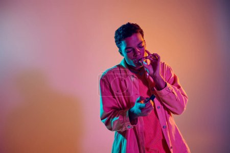Téléchargez les photos : Cool homme afro-américain allumant une cigarette tout en tenant plus léger sous les néons bleus lumières sur rose - en image libre de droit