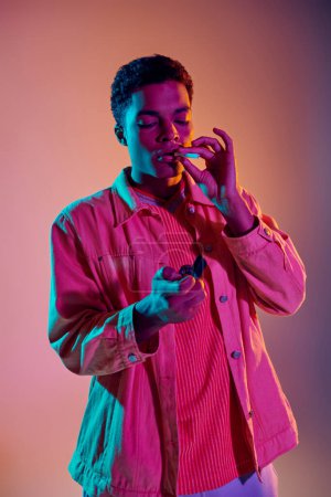 Téléchargez les photos : Afro-américain allumant une cigarette tout en tenant un briquet sous des néons bleus sur rose - en image libre de droit