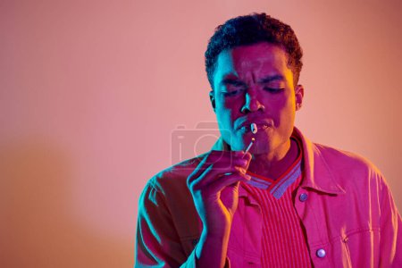 chico afroamericano enfocado iluminación cigarrillo con partido bajo luces de neón azul sobre fondo rosa
