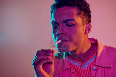 focused african american man lighting cigarette with match under blue neon lights on pink background