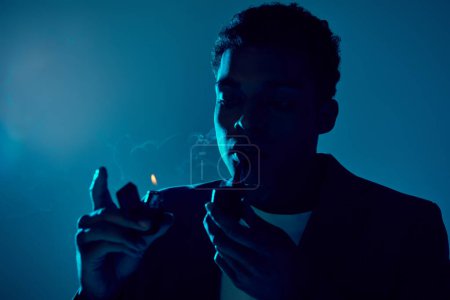 curly african american man holding lighter and smoking pipe on dark blue background with lighting