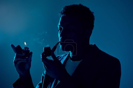 young african american man holding lighter and smoking pipe on dark blue background with lighting