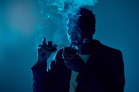 curly african american man holding lighter and pipe while exhaling smoke on dark blue background