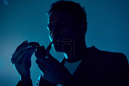 curly african american man holding lighter while smoking pipe on dark blue backdrop with lighting