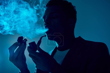 african american guy holding lighter and pipe while exhaling smoke on dark blue background