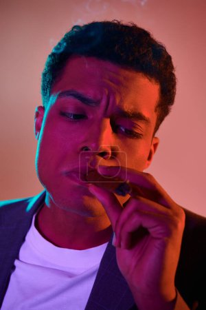 Téléchargez les photos : Portrait de jeune homme afro-américain avec cigare dans la bouche sur fond rose avec éclairage bleu - en image libre de droit