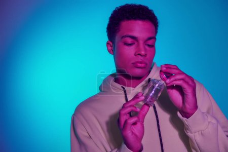 young african american man in hoodie holding jar with medical cannabis on blue background