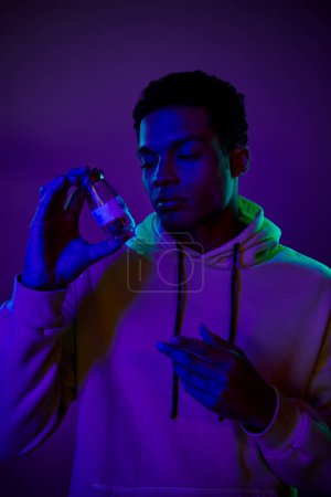 young african american man in hoodie holding jar with medical cannabis on dark blue background