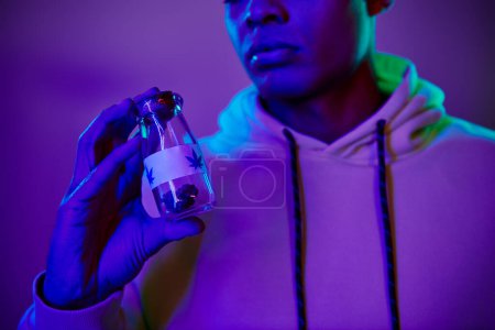 cropped african american man in hoodie holding jar with medical cannabis on dark blue background