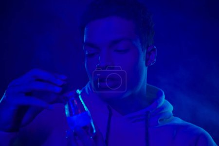 african american man in hoodie looking at jar with medical cannabis on dark blue around smoke