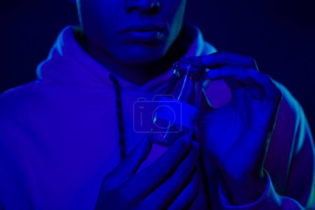 cropped african american man in hoodie holding jar with medical cannabis on dark blue around smoke