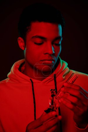 handsome african american man in hoodie looking at glass bong on dark background with red lighting
