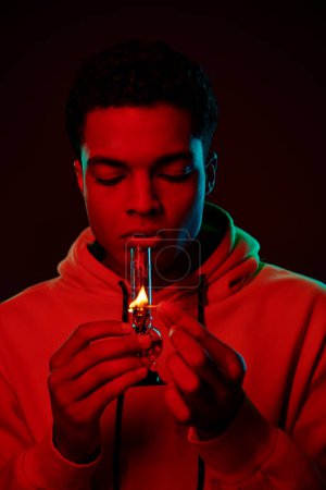 young african american man in hoodie lighting glass bong on dark background with red lighting
