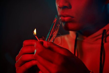 cropped african american man in hoodie lighting glass bong on dark background with red lighting