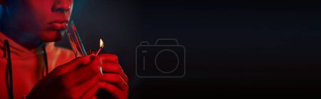 cropped banner of black man in hoodie lighting glass bong on dark background with red lighting