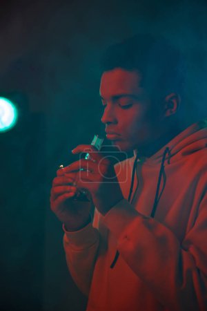 smoke around african american guy in hoodie holding glass bong on dark background, medical cannabis