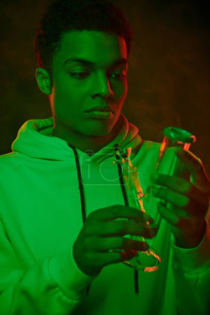 junger afrikanisch-amerikanischer Mann in Kapuzenpulli mit Blick auf Glasbong auf dunklem Hintergrund mit grüner Beleuchtung