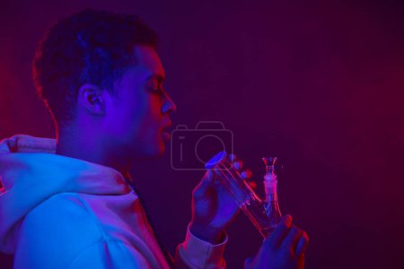 Téléchargez les photos : Jeune homme afro-américain en sweat à capuche regardant bong en verre sur fond violet foncé avec éclairage - en image libre de droit