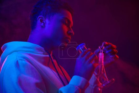 Téléchargez les photos : Jeune homme afro-américain à capuche éclairage verre bong sur fond bleu foncé avec lumière rouge - en image libre de droit
