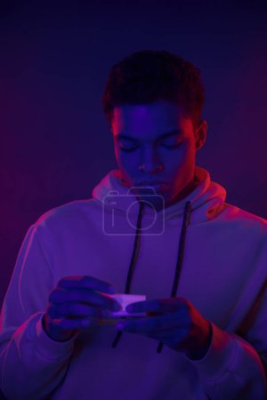 Téléchargez les photos : Jeune homme afro-américain rouler cigarette tout en se tenant debout sur fond bleu foncé avec lumière au néon - en image libre de droit