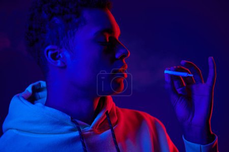 Téléchargez les photos : Afro-américain tenant roulé cigarette tandis que debout sur fond bleu foncé avec néon lumière - en image libre de droit