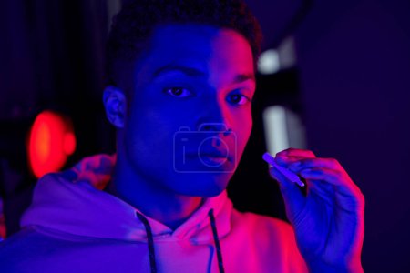 hombre afroamericano fresco sosteniendo el cigarrillo enrollado sobre fondo azul oscuro con luz roja