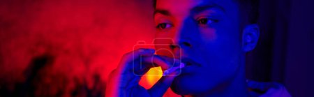 Téléchargez les photos : Afro-américain tenant la cigarette roulée près de la bouche sur bleu foncé avec lumière rouge, bannière fumeur - en image libre de droit