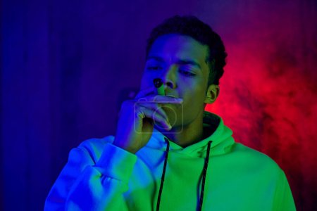 serious african american young man in hoodie smoking cigar on dark blue background with red light
