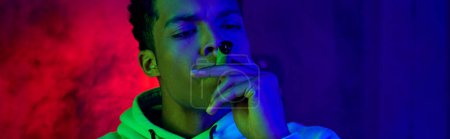 estandarte de afroamericano joven en sudadera con capucha fumar cigarro sobre fondo azul oscuro con luz roja