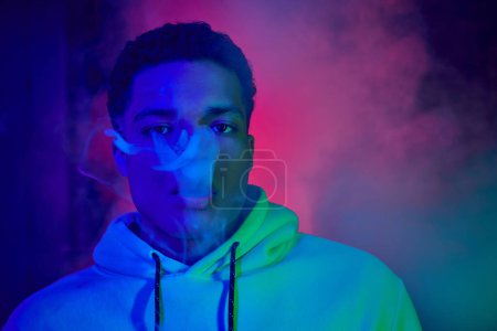 Photo for Cool african american man exhaling smoke on dark background with blue and red light, smoker - Royalty Free Image