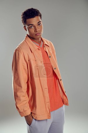 junger afrikanisch-amerikanischer Mann in pfirsichfarbenem Hemd blickt in die Kamera vor grauem Hintergrund, Hand in Tasche
