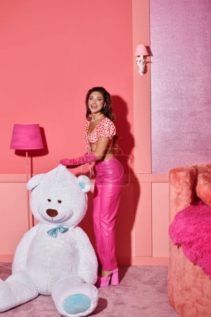 fröhliche junge Frau in rosa bauchfreiem Top und Hose lacht in der Nähe eines riesigen Teddybären in einem lebhaften Raum