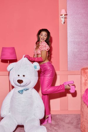 excited and young woman in pink crop top with pants posing on high heels near giant teddy bear