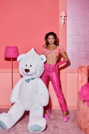 Glamouröse junge Frau in rosa Crop Top und Hose posiert mit der Hand auf der Hüfte in der Nähe von Riesen-Teddybär