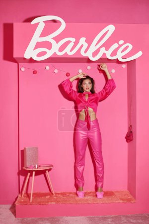Téléchargez les photos : KYIV, UKRAINE - 14 JUILLET 2023 : jeune femme glamour en haut de la culture et pantalon rose debout avec les mains levées près des lettres Barbie - en image libre de droit