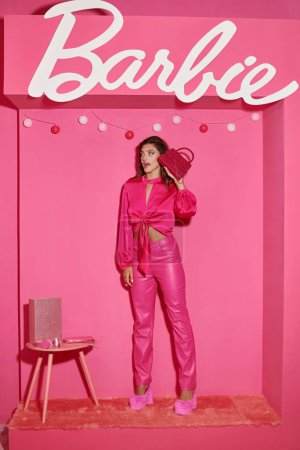 Téléchargez les photos : KYIV, UKRAINE - 14 JUILLET 2023 : étonné et poupée comme une femme en haut de la culture et pantalon rose debout avec sac à main chic près signe Barbie - en image libre de droit