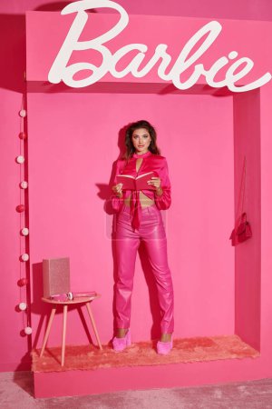Téléchargez les photos : KYIV, UKRAINE - 14 JUILLET 2023 : belle poupée comme une femme en haut de la culture et pantalon rose debout avec livre près de signe Barbie, glamour - en image libre de droit