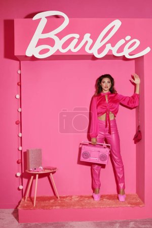 Photo for KYIV, UKRAINE - JULY 14, 2023:beautiful young woman in pink attire holding retro boombox and waving hand near Barbie sign - Royalty Free Image