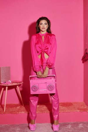schöne junge Frau in lebendiger Kleidung mit Retro-Boombox und Blick in die Kamera auf rosa Hintergrund