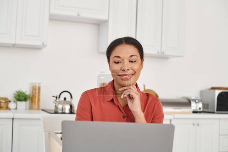 Trabajo remoto de joven nutricionista afroamericana alegre usando el ordenador portátil en su cocina, mujer