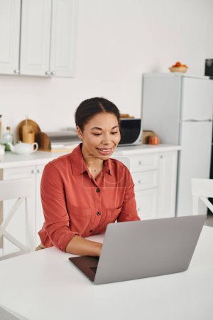 cheerful young african american nutritionist using laptop while working remotely from her kitchen