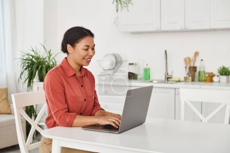 happy african american nutritionist using laptop while working remotely from her kitchen