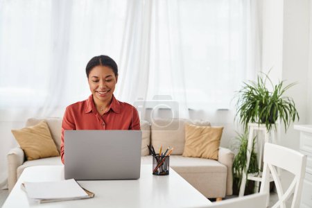 Foto de Mujer afroamericana feliz usando el ordenador portátil mientras trabaja desde su cocina moderna, nutricionista - Imagen libre de derechos
