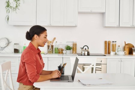 Photo for Cheerful african american woman using laptop while working from her modern kitchen, nutritionist - Royalty Free Image