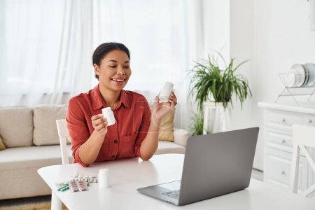 Glückliche Ernährungsberaterin zeigt Flaschen mit verschiedenen Medikamenten bei Online-Beratung am Laptop