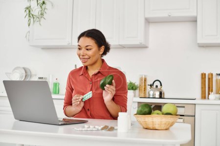 Photo for African american nutritionist holding avocado and supplements while giving diet advice online - Royalty Free Image