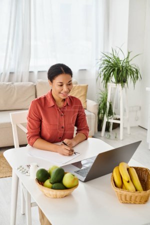 happy african american nutritionist taking notes while looking at laptop during online consultation